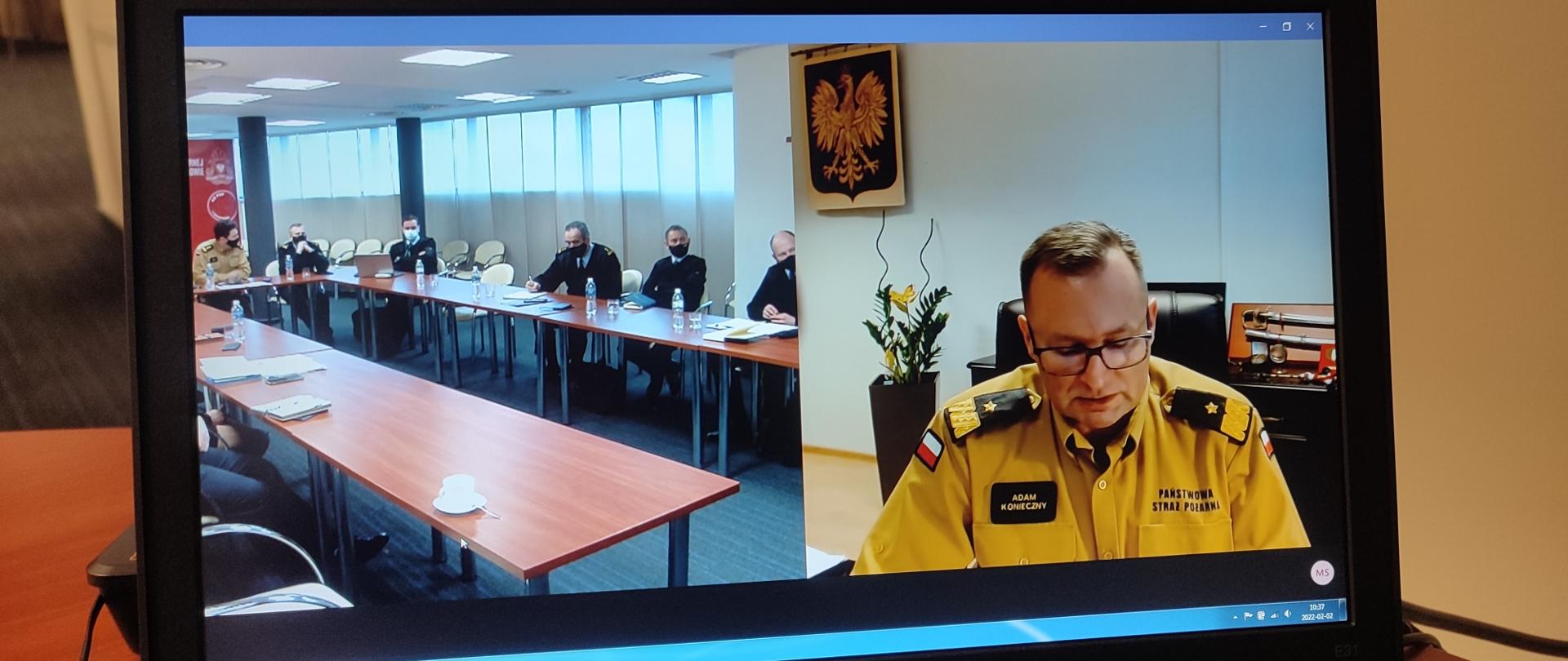Zdjęcie przedstawia ekran laptopa. Na lewej połowie ekranu widoczny widok sali konferencyjnej Centralnej Szkoły PSP, w której siedzą uczestnicy narady. Po prawej stronie ekranu widoczny Zastępca Komendanta Głównego PSP w swoim gabinecie