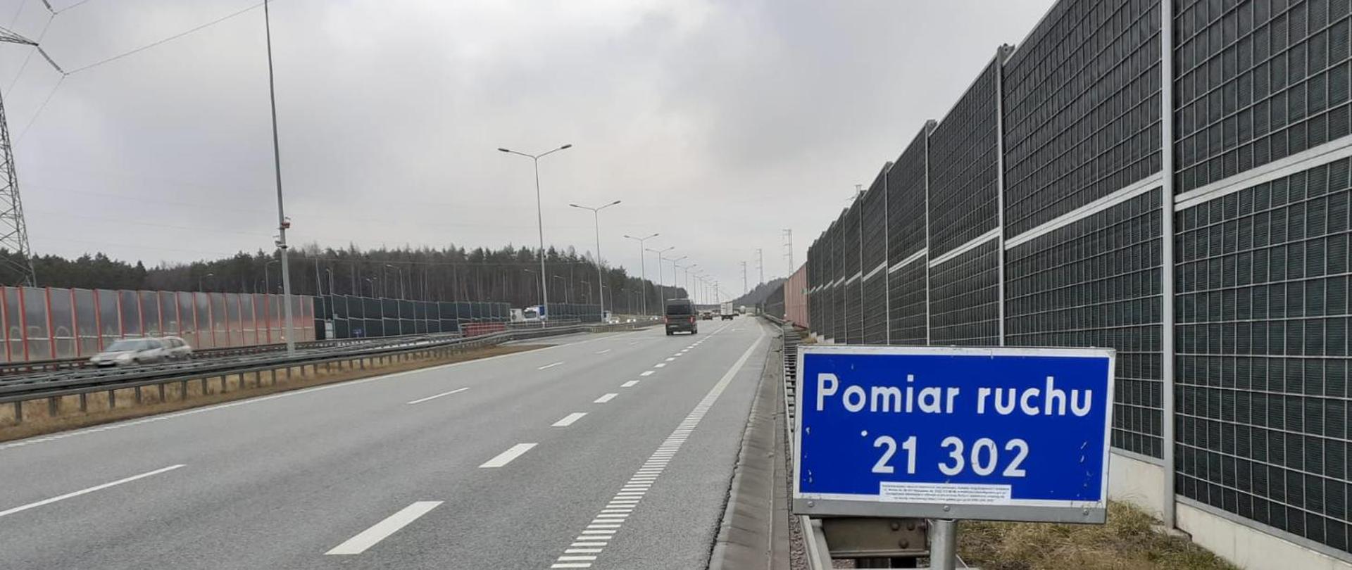 Generalny Pomiar Ruchu - przy jednej jezdni drogi dwujezdniowej niebieska tabliczka z cyfrowym oznaczeniem punktu pomiarowego i napisem "pomiar ruchu". Wzdłuż jezdni bariery ochronne i ekrany akustyczne po zewnętrznej stronie. 