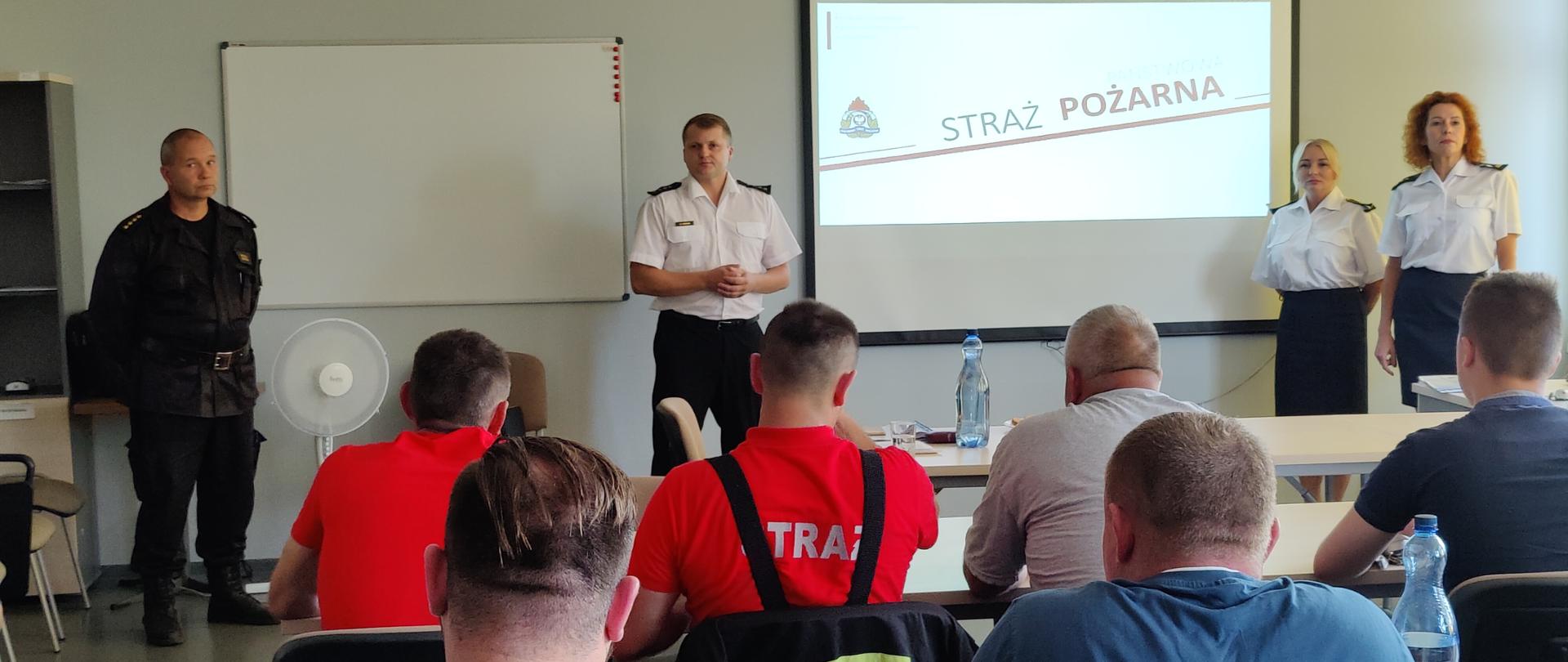Strażacy PSP i OSP siedzą w ławkach przy sali wykładowej, przy wyświetlonej prezentacji stoją dwie strażaczki ubrane w białe koszule i granatowe spódnice, z drugiej strony dwóch strażaków - jeden w białej koszuli i grantowych spodniach, drugi w ubraniu sztabowym 