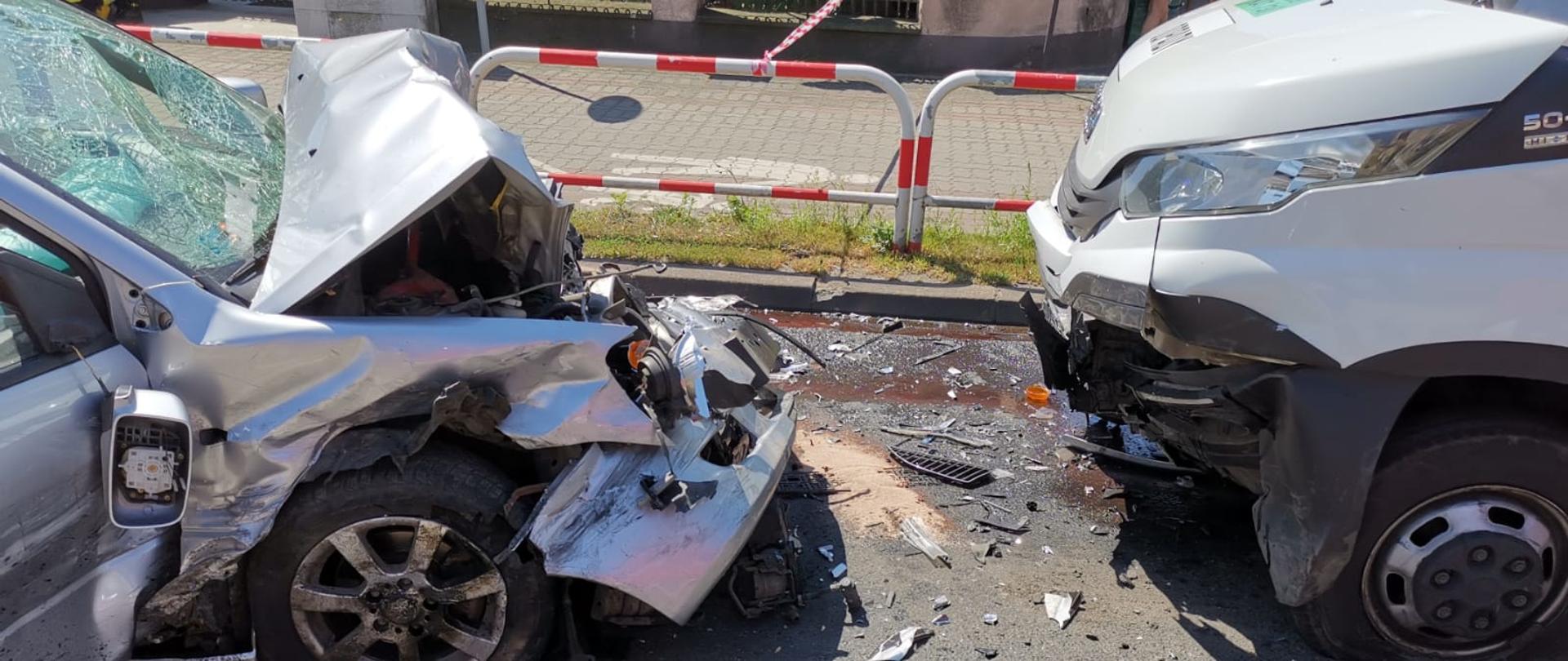 Przód samochodu osobowego i busa, które się zderzyły czołowo. Samochód osobowy ma całkowicie wgnieciony przód. Na jezdni porozlewane płyny eksploatacyjne z pojazdów.