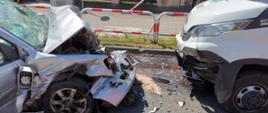 Przód samochodu osobowego i busa, które się zderzyły czołowo. Samochód osobowy ma całkowicie wgnieciony przód. Na jezdni porozlewane płyny eksploatacyjne z pojazdów.