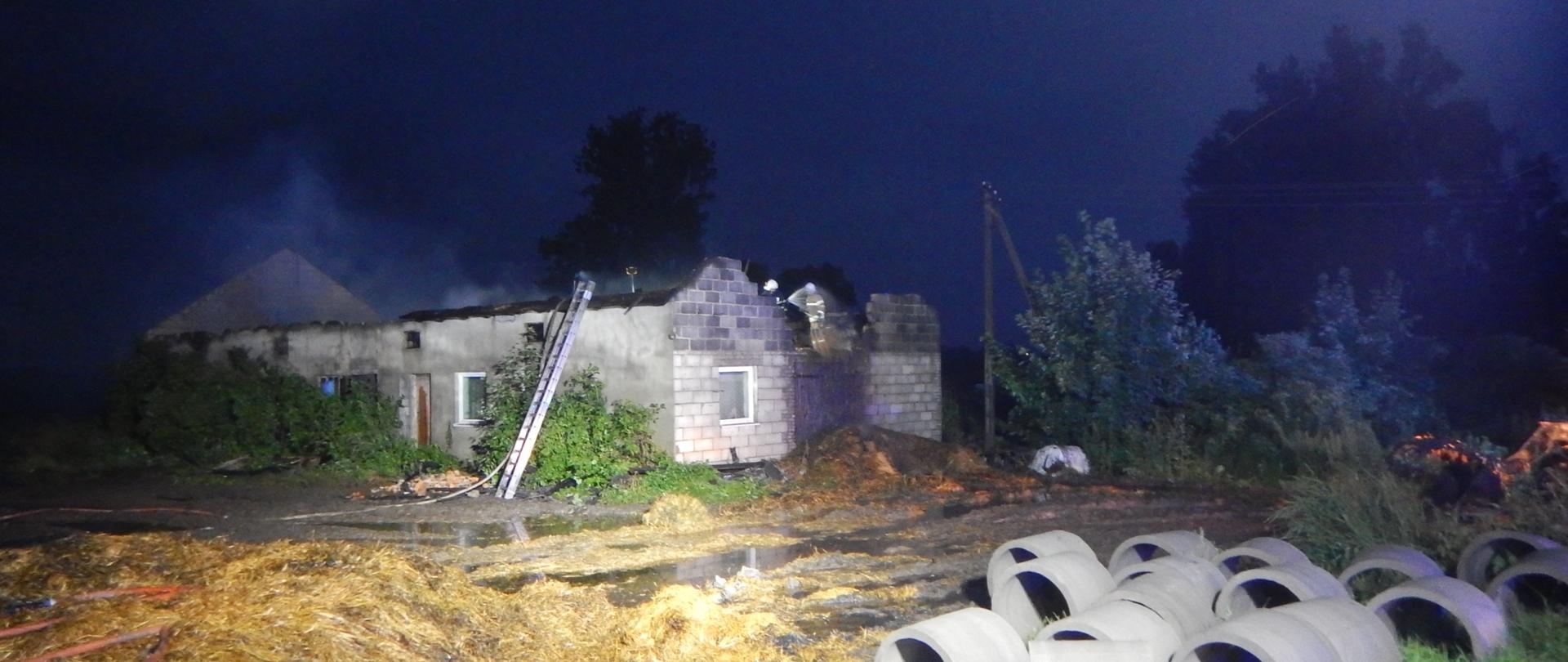 Zdjęcie przedstawia budynek obory, który uległ spaleniu. Na zdjęciu widać strażaków pracujących przy rozbiórce konstrukcji.