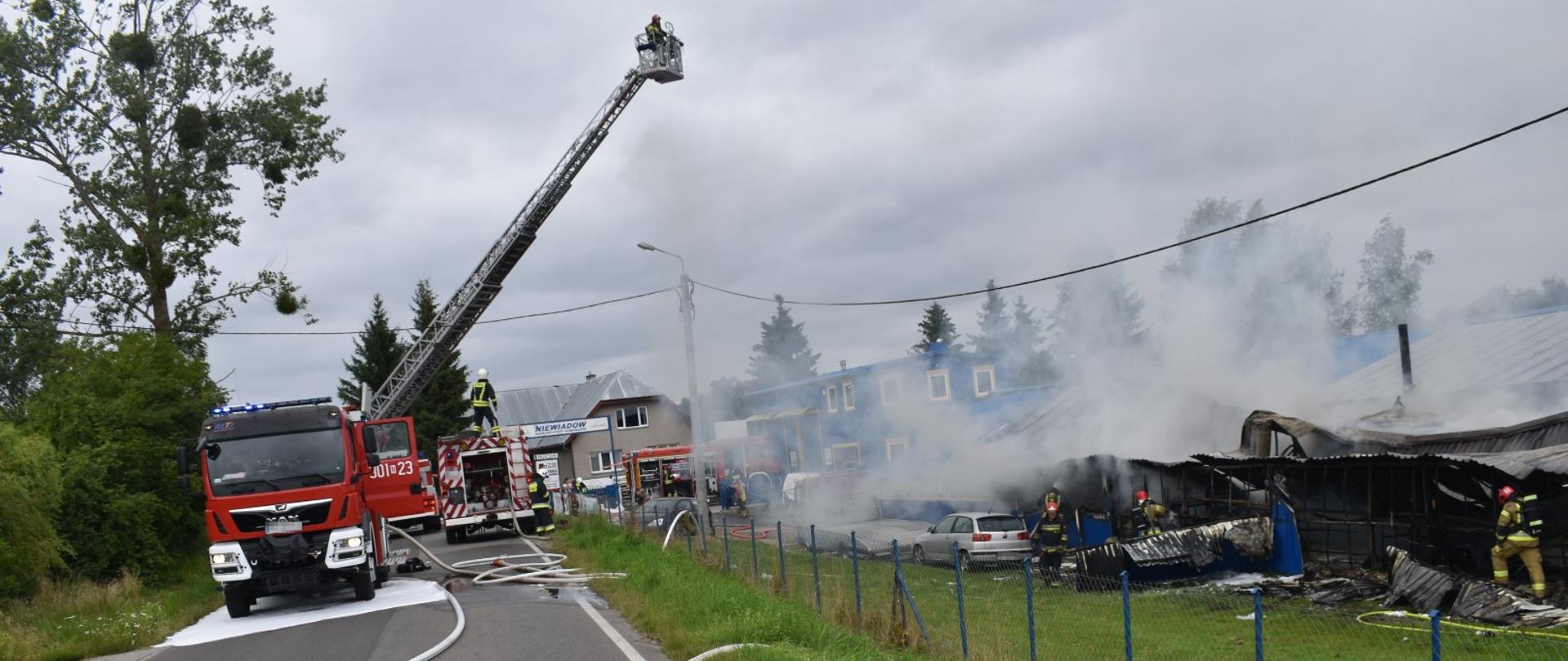 Zdjęcie ukazuje zastępy Państwowej i Ochotniczej Straży Pożarnej podczas gaszenia pożaru hali z przyczepami kempingowymi w miejscowości Bartąg