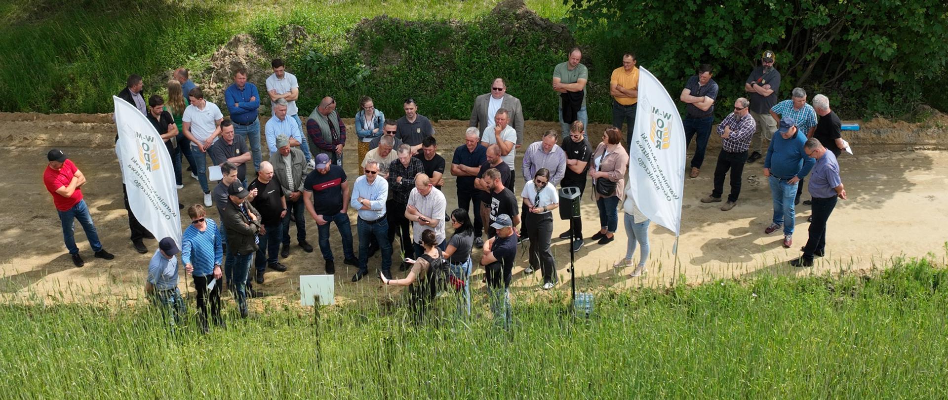 Grupa ludzi stoi na polu, otoczona zielenią i naturalnym krajobrazem.