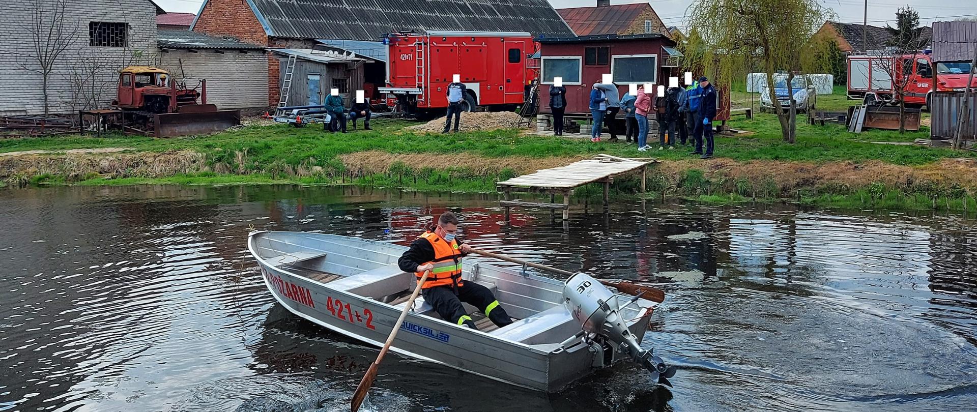 Na zdjęciu strażak PSP w kamizelce asekuracyjnej wiosłuje łodzią po stawie. W tle samochody pożarnicze oraz osoby postronne.