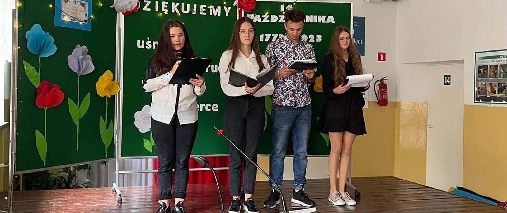 Grupa czworga uczniów na scenie czyta teksty z czarnych folderów. W tle dekoracja z kolorowymi papierowymi kwiatami i napisem „14 października”. Na pierwszym planie biała kolumna głośnikowa.