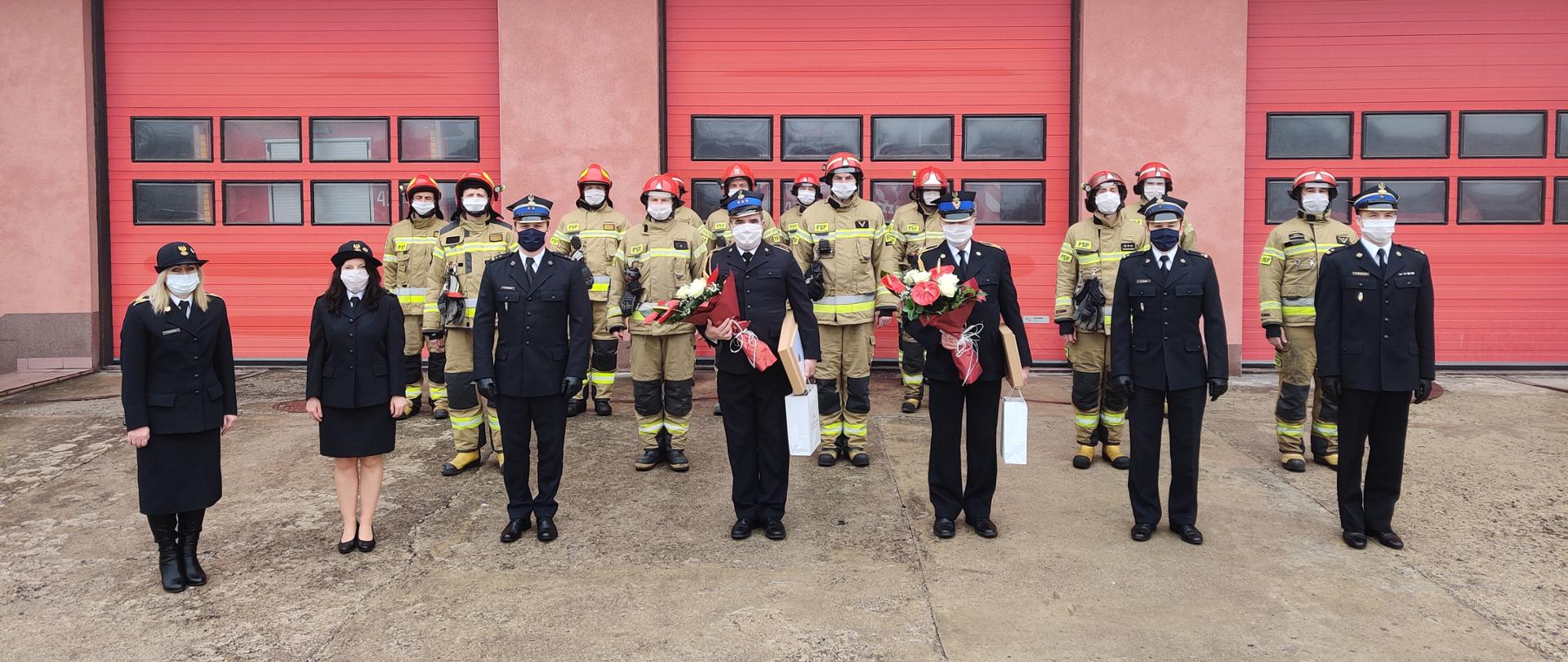 W szyku stoją strażacy w żółtych i granatowych mundurach. W środku dwóch strażaków trzyma kwiaty.