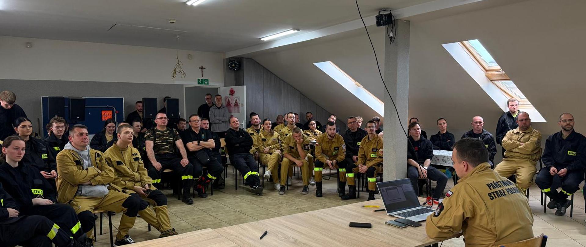 Zdjęcie przedstawia grupę strażaków słuchających wykładowcy za biurkiem.