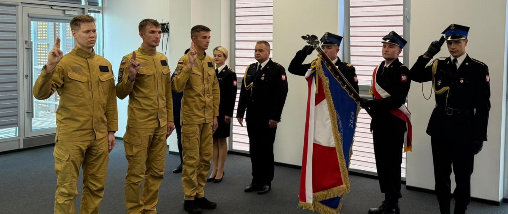 Na zdjęciu widocznych jest trzech nowo przyjętych strażaków składających ślubowanie strażackie. W tle widoczny jest poczet sztandarowy jednostki podczas uroczystości