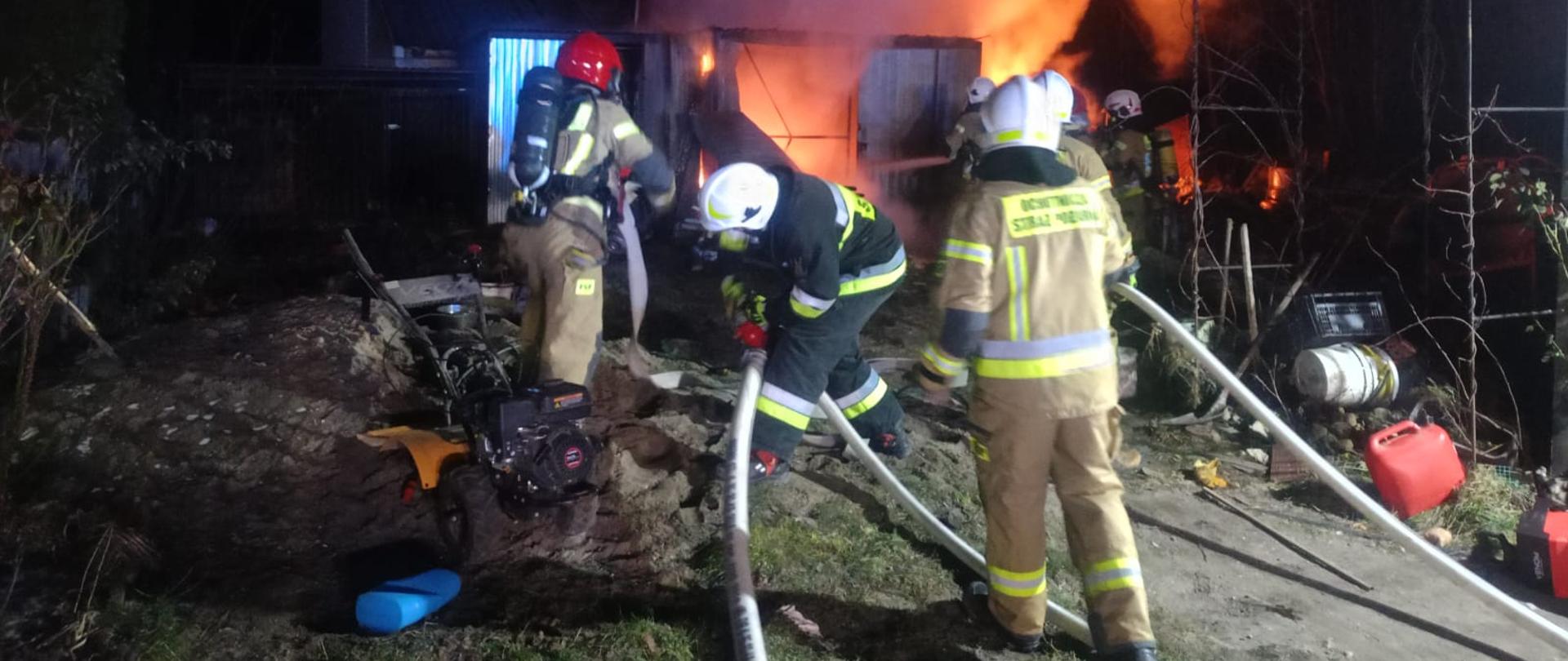 na 1 planie strażacy stoją tyłem, ciągną linię wężową, na wprost palący się garaż. Po prawej stronie widać częściowo wyniesione wyposażenie palącego się garażu 