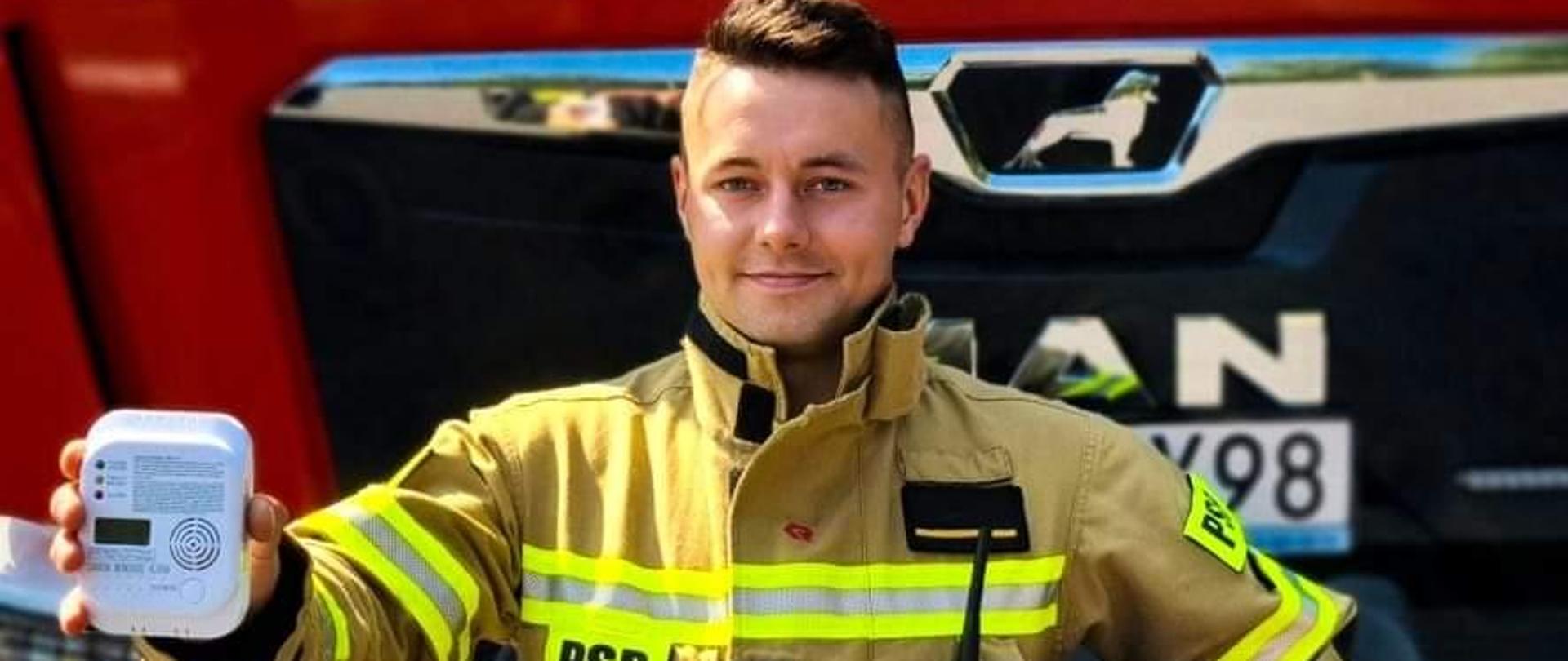 Funkcjonariusz Państwowej Straży Pożarnej w ubraniu specjalnym pokazujący czujkę na tle przodu samochodu ratowniczo gaśniczego. 