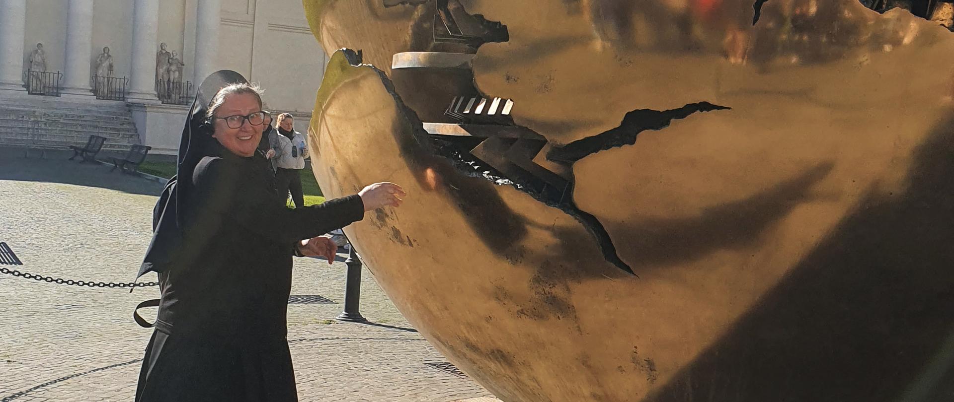 Fotografia zostało wykonana na Dziedzińcu Pinii w Watykanie. Głównym elementem tego zdjęcia jest rzeźba - eksponat „Kula w kuli” autorstwa Arnaldo Pomodoro, która została zrealizowana w 1990 r. Kiedy wieje wiatr, sfera porusza się. Obok rzeźby stoi Siostra Grażyna Powierża, która stara się poruszyć, wprawić w ruch wirowy potężną brązową kulę. 