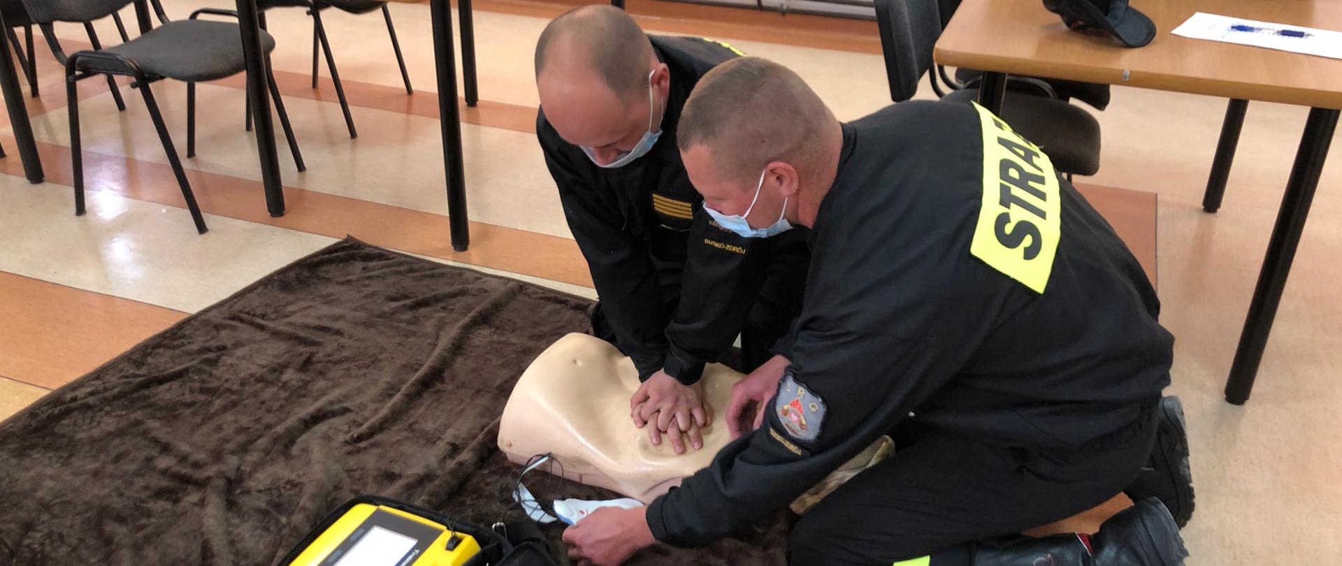 Na pierwszym zdjęciu strażacy-ratownicy wykonują symulację resuscytacji krążeniowo-oddechowej u osoby dorosłej z użyciem defibrylatora AED. Jeden z ratowników wykonuje 30 uciśnięć klatki piersiowej, natomiast drugi wykonuje 2 oddechy ratownicze za pomocą worka samorozprężalnego oraz przykleja elektrody defibrylatora na klatce piersiowej poszkodowanego.