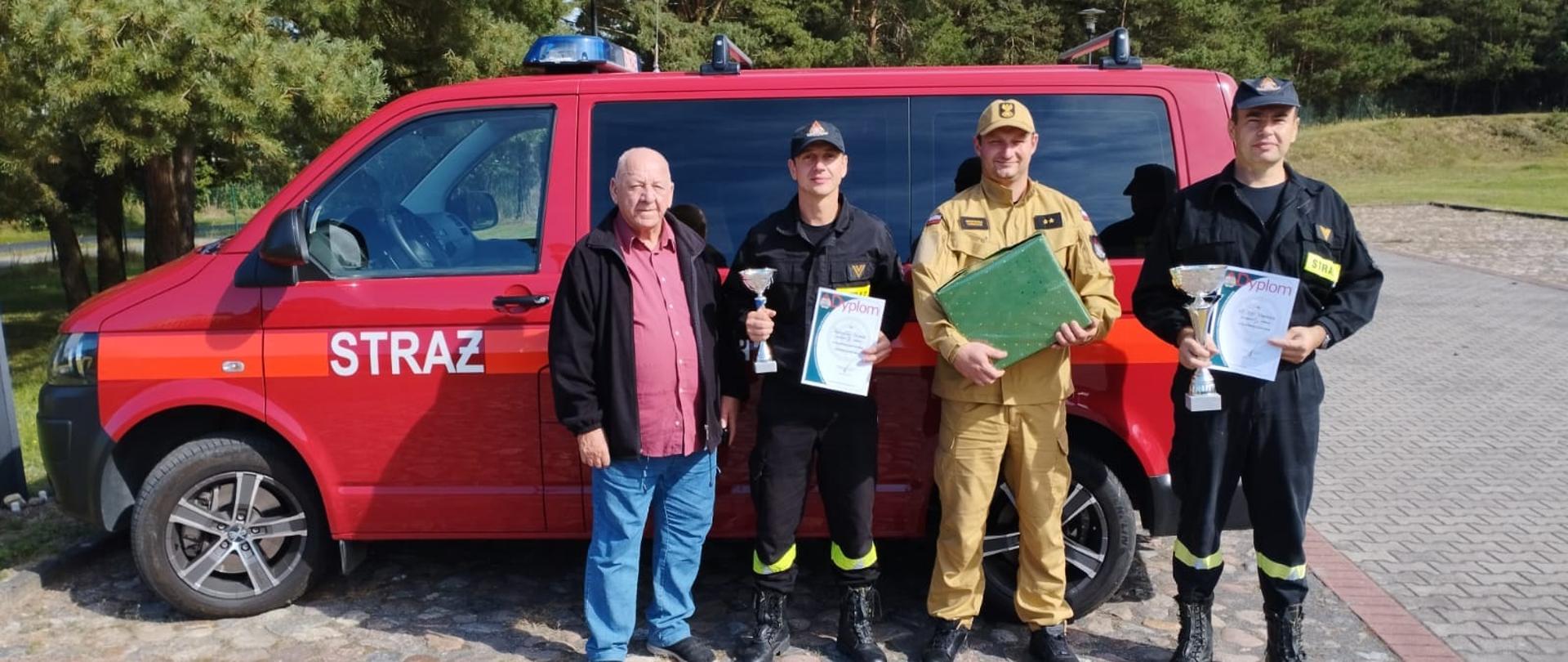 Zdjęcie przedstawia trzech strażaków z dyplomami oraz z trenerem na tle samochodu pożarniczego