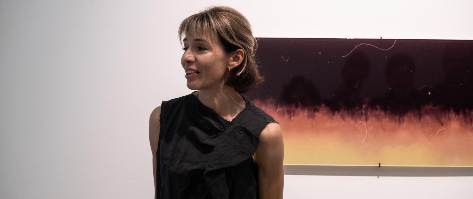 Polska fotografka Katerina Kouzmitcheva stoi w przestrzeni galerii przed jedną z prezentowanych fotografii. Uśmiecha się i zwraca w stronę uczestników wernisażu, opowiadając o swojej twórczości. Za nią widoczna jest wielkoformatowa praca z cyklu prezentowanego na wystawie „Przepraszam za brak kontaktu”.