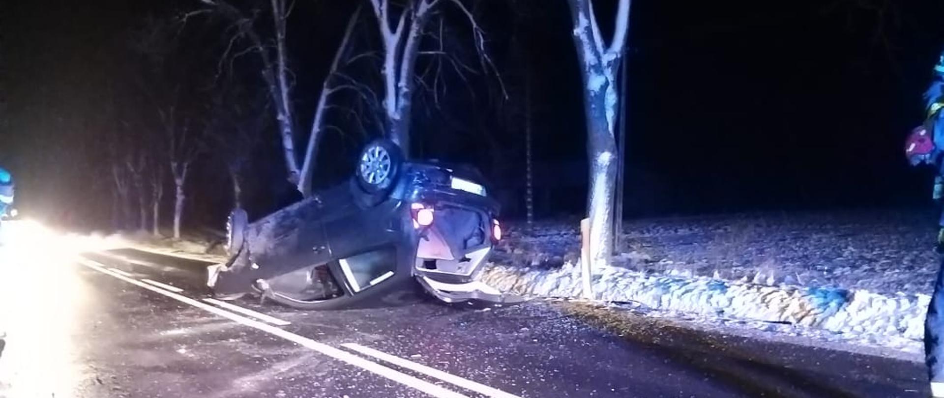 Na zdjęciu samochód osobowy VW Polo leżący na dachu pojazdu w poprzek drogi wojewódzkiej. Oświetlone miejsce zdarzenia z uwagi na godzinę poranną.