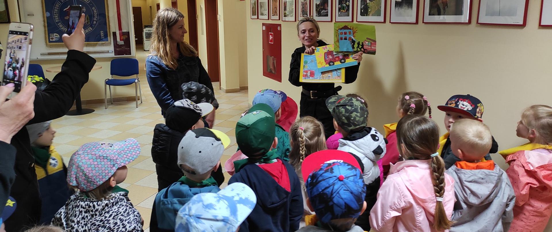 Na zdjęciu jasny korytarz z holem, na którym stoi duża grupa dzieci. Przed dziećmi kobieta w czarnym mundurze trzyma w dłoniach trzy obrazki z dziecięcymi rysunkami przedstawiającymi pracę strażaków. Obok po lewej stronie obrócona prawym bokiem wychowawczyni. W tle ściana z fotografiami w ramkach przedstawiająca różnego rodzaju działania ratownicze.