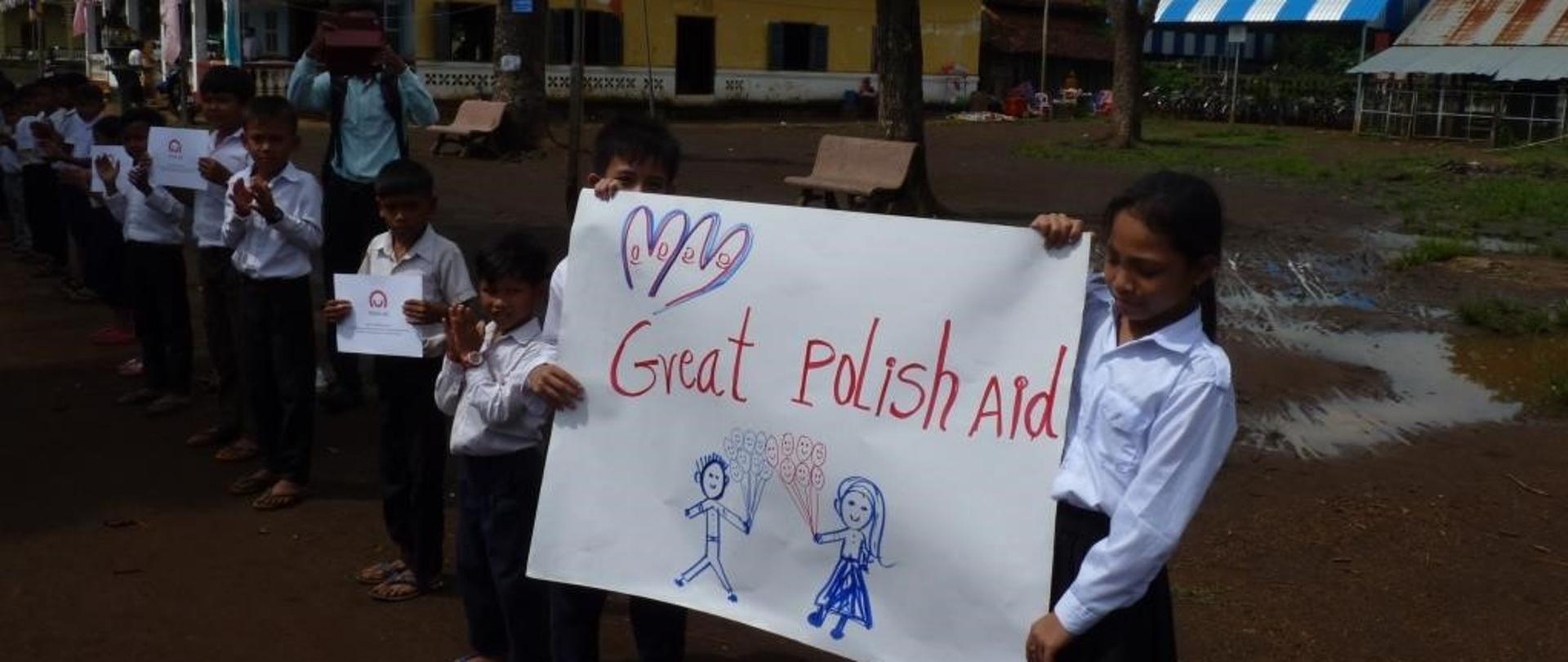 dzieci ubrane w uroczyste stroje galowe trzymają napis Great Polish Aid