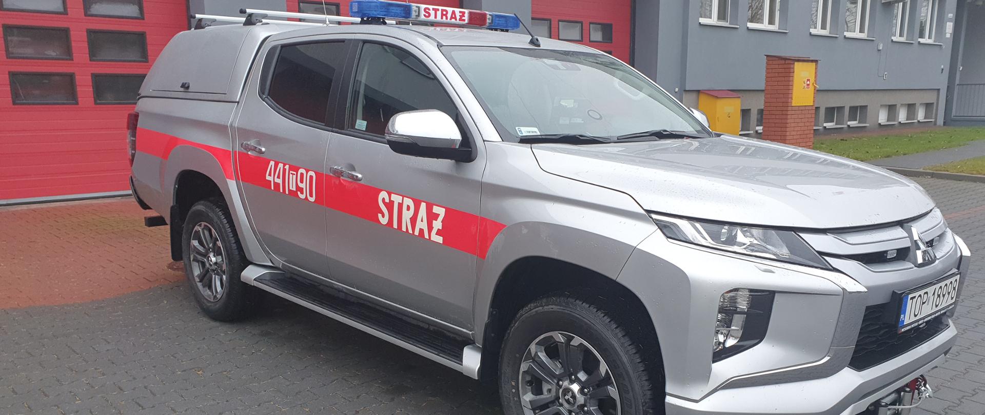 Zdjęcie przedstawia srebrny samochód rozpoznawczo ratowniczy stojący na parkingu. W tle budynek PSP Opatów