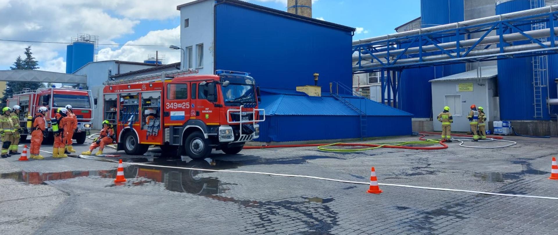 Zdjęcie przedstawia plac zakładowy SM Mlekovita Kurpie w Baranowie podczas ćwiczeń ratowniczo-gaśniczych. Na pierwszym planie, na mokrej kostce brukowej, stoją dwa czerwone samochody strażackie z otwartymi skrytkami sprzętowymi. Wokół pojazdów oraz w głębi placu, przy niebieskich budynkach przemysłowych i silosach, widoczni są strażacy w pełnym umundurowaniu bojowym i kombinezonach ochronnych chemicznych. Teren zabezpieczony za pomocą stożków ostrzegawczych. Rozwinięte węże gaśnicze .