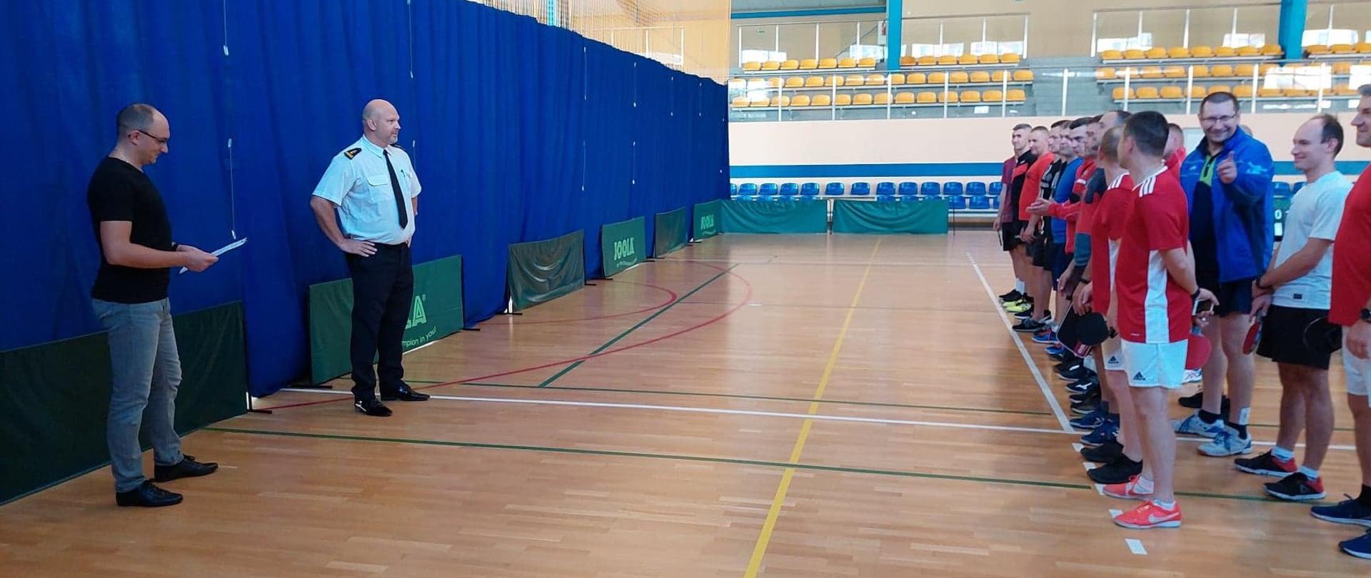 Startujący zawodnicy w strojach sportowych stoją na hali w dwuszeregu naprzeciwko Komendanta Powiatowego PSP oraz Sędziego Głównego.