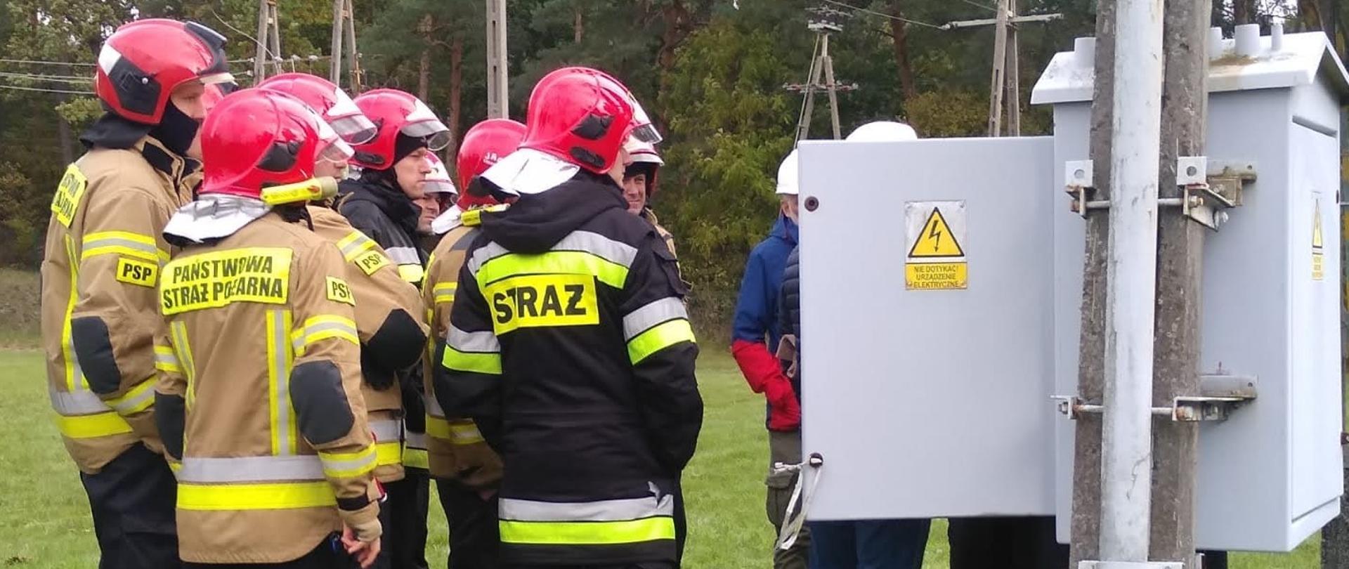 Strażacy Państwowej Straży Pożarnej stoją obok urządzenia pod napięciem, osoby z firmy Energa - Operator informują o zagrożeniach.