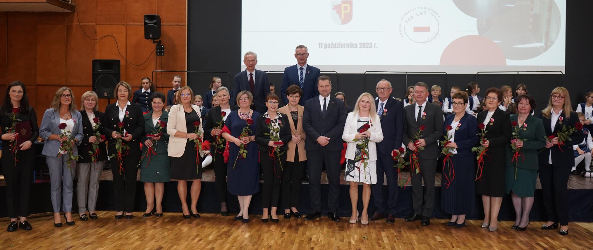 W sali na wyłożonej klepką podłodze pod ścianą z wielkim ekranem pokazującym napis Wielkie otwarcie stoi duża grupa ludzi trzymających kwiaty.