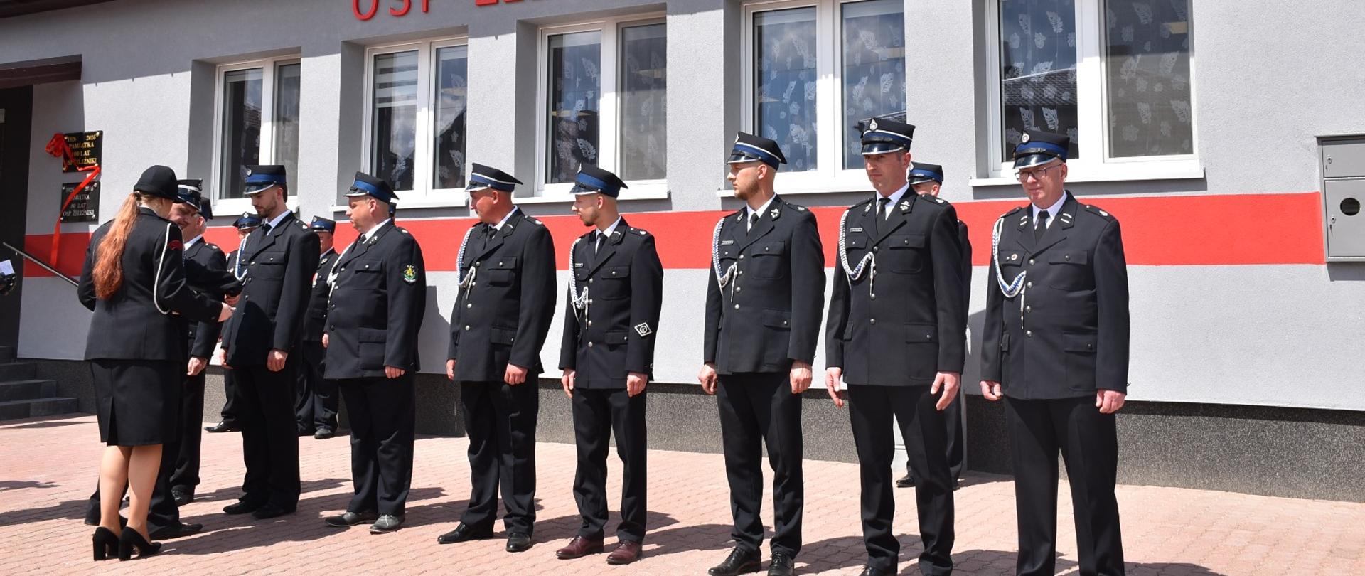 Oficjalne rozpoczęcie uroczystości przed odnowionym budynkiem remizy Ochotniczej Straży Pożarnej w Żeleźnikach (rok założenia 1926). Na placu w równym szyku stoją druhowie OSP w pełnym umundurowaniu galowym. Po lewej stronie widoczny jest moment składania raportu lub wręczania wyróżnień.