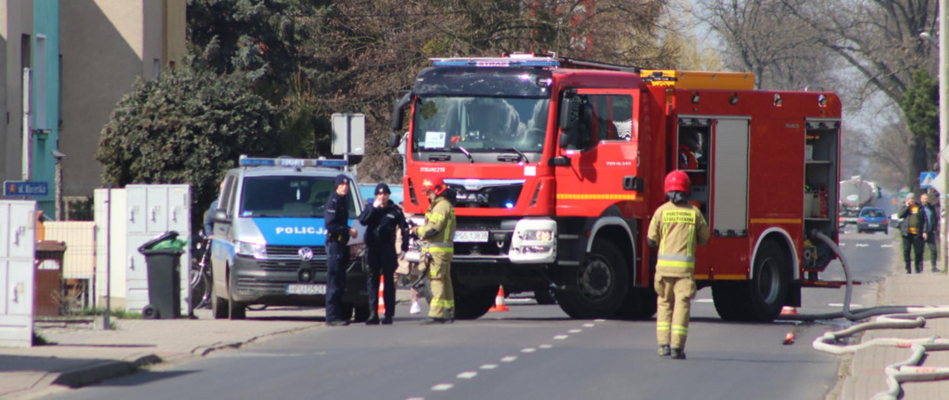 Zdjęcie przedstawia zastęp straży oraz Policje, stojące na drodze podczas działań związanych z gaszeniem pożaru butli z acetylenem.