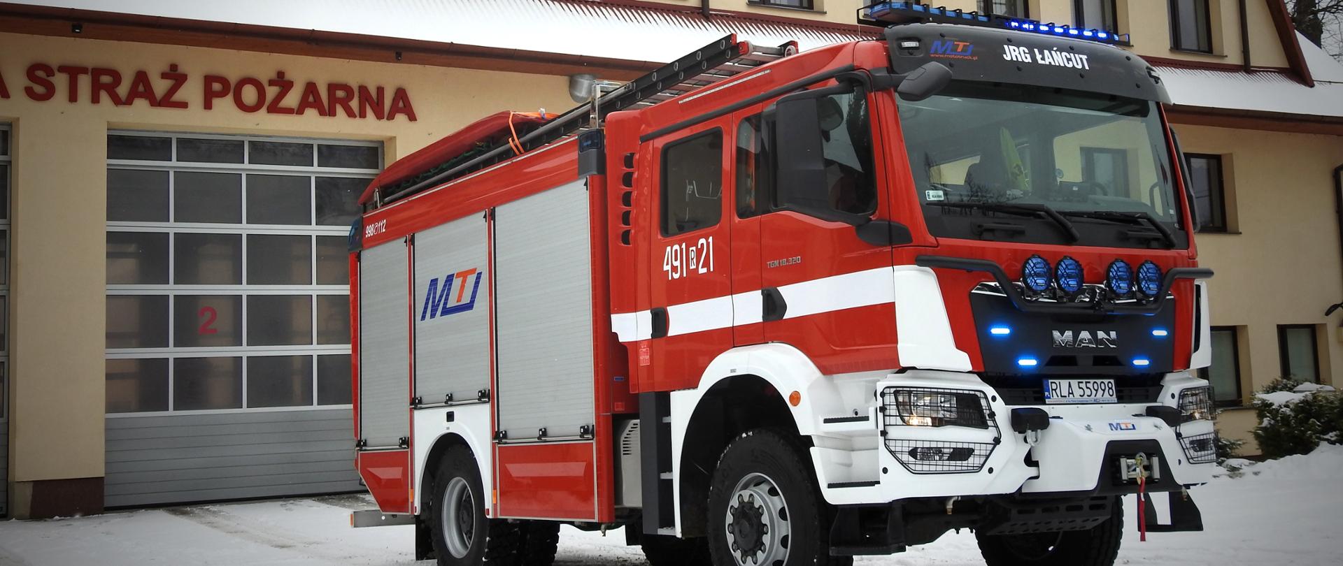Na zdjęciu nowo zakupiony średni samochód ratowniczo gaśniczy na podwoziu Man. Pojazd stoi przed budynkiem komendy widoczny z przodu i prawej strony. Samochód ma włączone oświetlenie alarmowe w kolorze niebieskim.