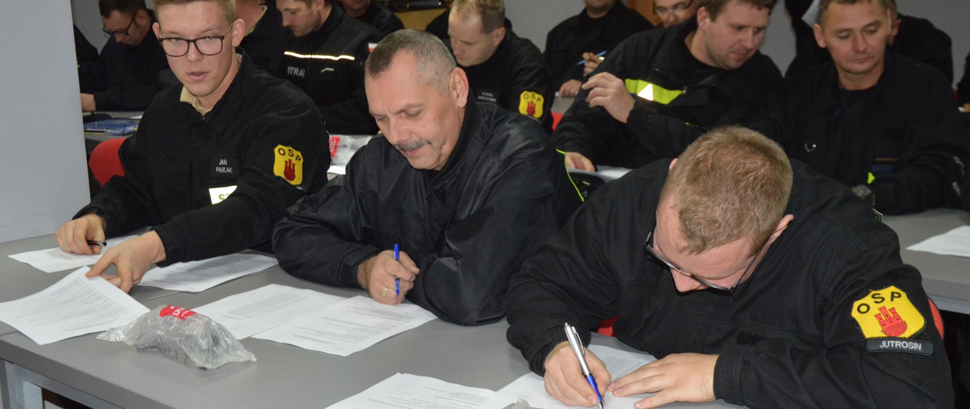 Recertyfikacja kpp. Świetlica w komendzie PSP w Rawiczu. Przy stołach siedzą druhowie OSP w umundurowaniu koszarowym, którzy uczestniczą w egzaminie recertyfikacyjnym. Zwróceni są w stronę okien. Strażacy rozwiązują test pisemny.