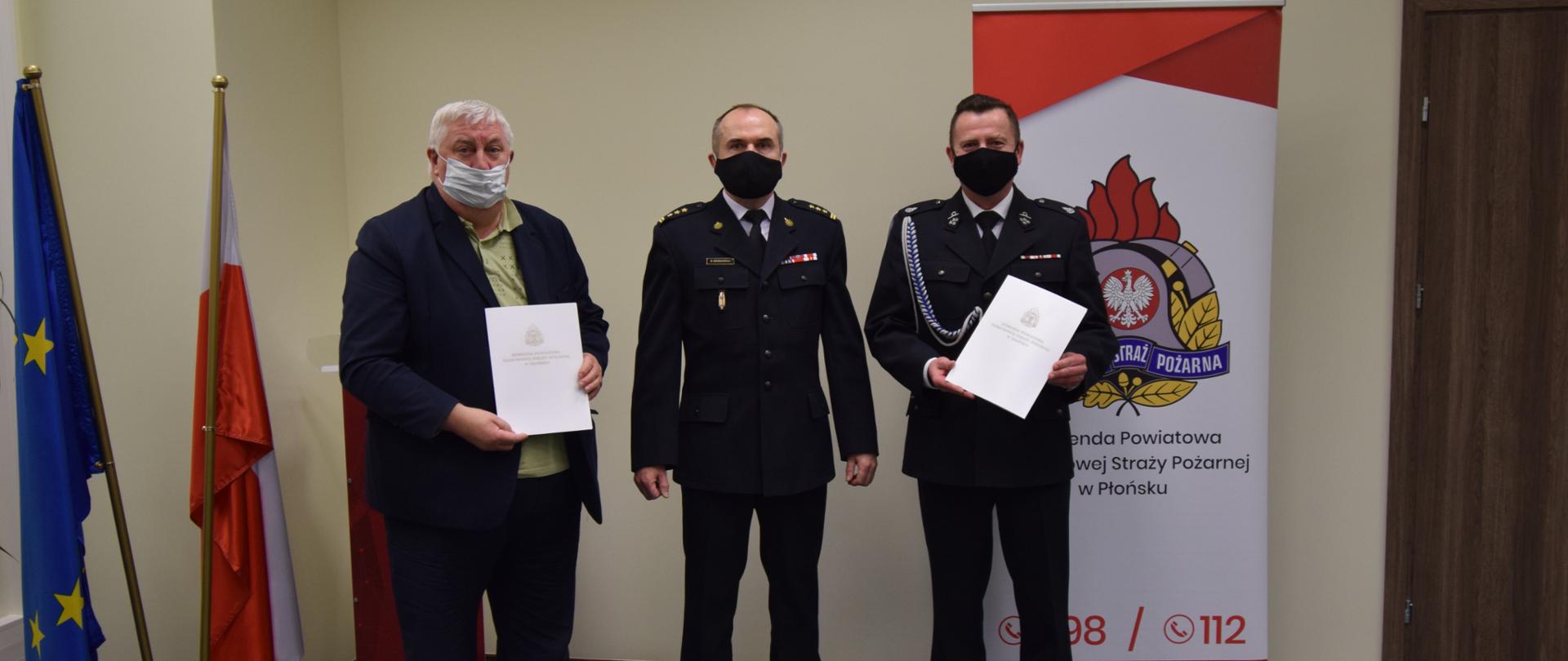 Na zdjęciu stoją trzy osoby z podpisanymi umowami dotyczącymi włączenia OSP Kownaty do KSRG. W tle po lewej stronie znajdują się flagi Polski i Unii Europejskiej. Po prawej stronie znajduje się logo PSP