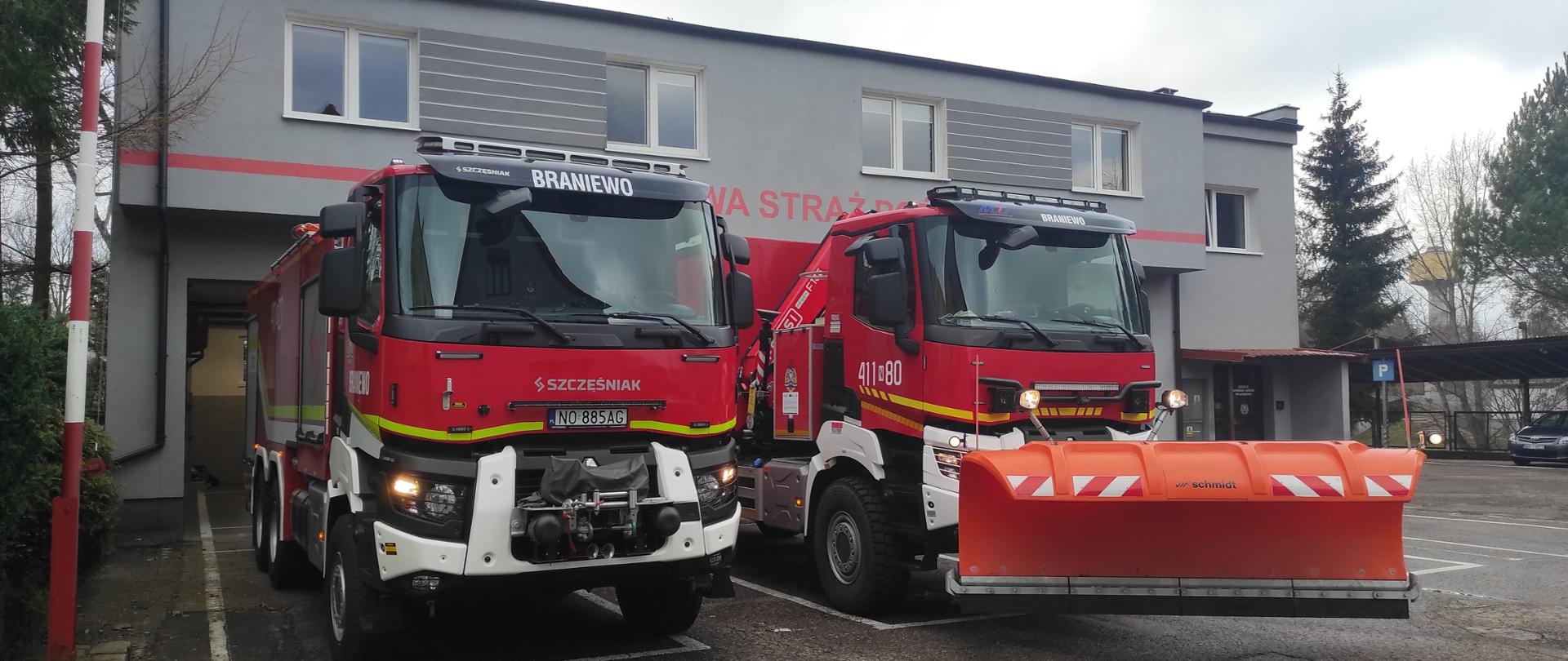 Na zdjęciu dwa duże czerwone samochody strażackie. Pojazd z prawej strony ma zaczepiony pomarańczowy pług do odśnieżania. Z tyłu szary budynek strażnicy.