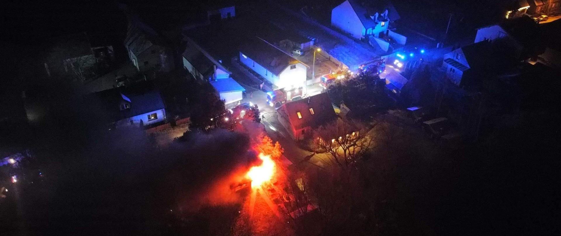 Zdjęcie z lotu ptaka, widać ogień przy budynku, w oddali kilka pojazdów na światłach błyskowych, jest noc