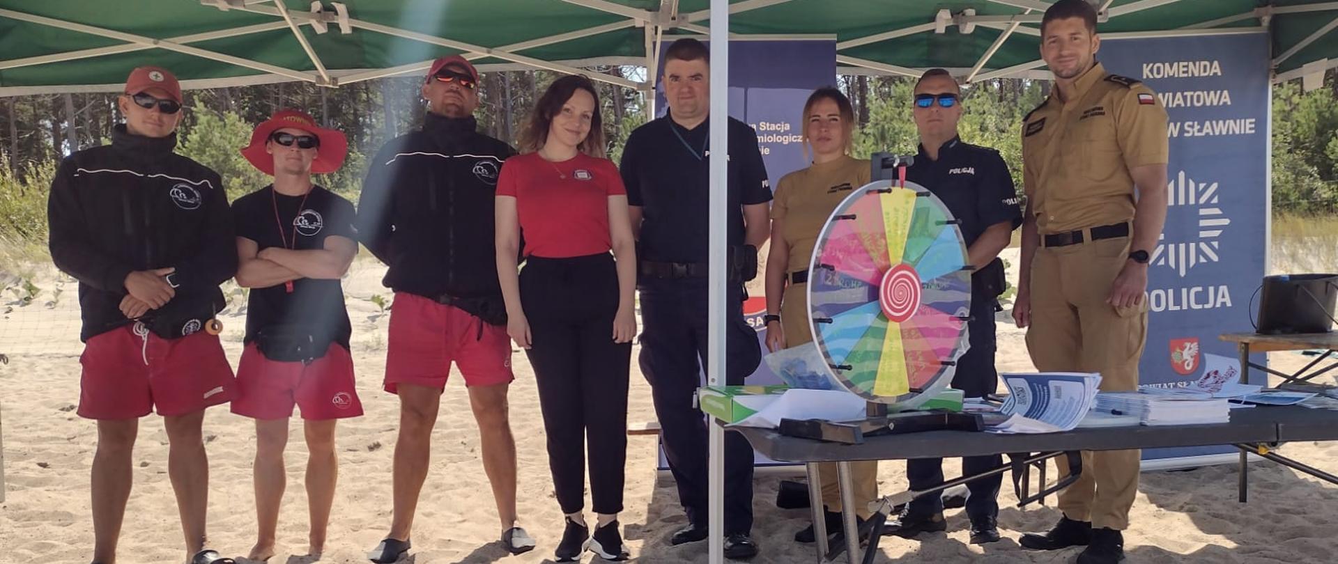 Grupa ratowników WOPR, funkcjonariuszy policji, przedstawicieli sanepidu i innych służb stoi razem pod zielonym namiotem na plaży, uśmiechając się do zdjęcia grupowego