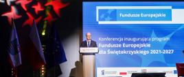 Ze sceny przemawia mężczyzna. W tle duży ekran z napisem "Konferencja inaugurująca program Fundusze Europejskie dla Świętokrzyskiego 2021-2027