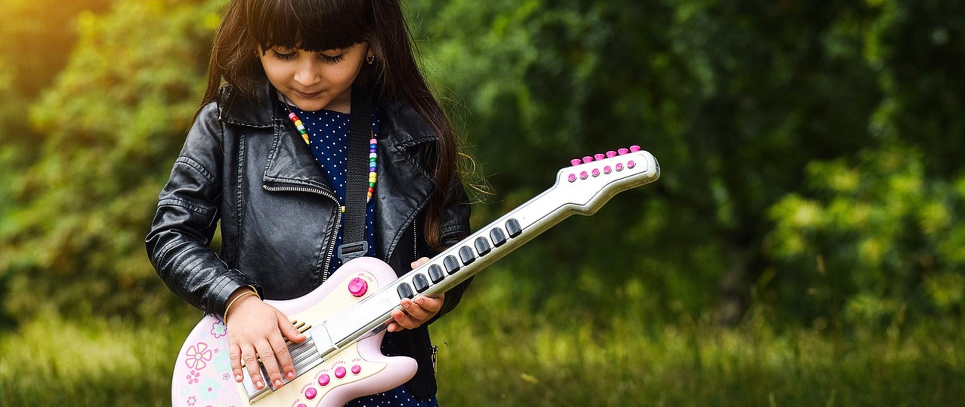Dziewczynka w skórzanej kurtce trzyma gitarę. Jest na tle zielonego lasu.