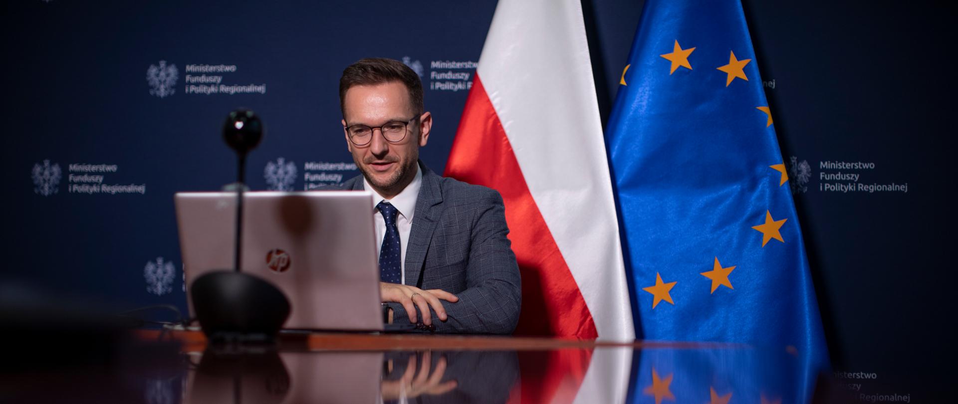 wiceminister Waldemar Buda siedzi przy komputerze za nim flagi Polski i Unii Europejskiej