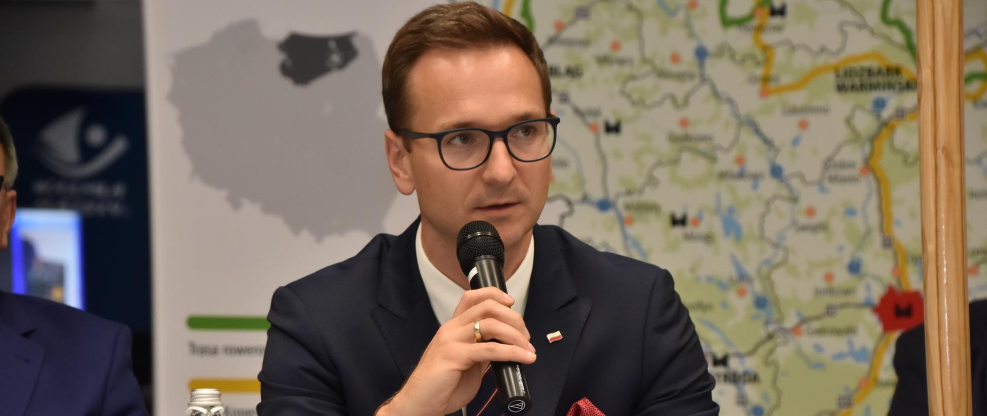 Wiceminister Waldemar Buda z mikrofonem siedzi przy stole, za nim mapa województwa warmińsko-mazurskiego.
