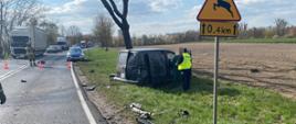 zdjęcie przedstawia odcinek drogi na nim porozbijane elementy pojazdów samochód zielony na poboczu drogi obok niego policjant i osoba postronna w oddali widać radiowóz policyjny oraz samochody stojące w korku przed nim strażak wokół pola oraz przy drodze drzewa