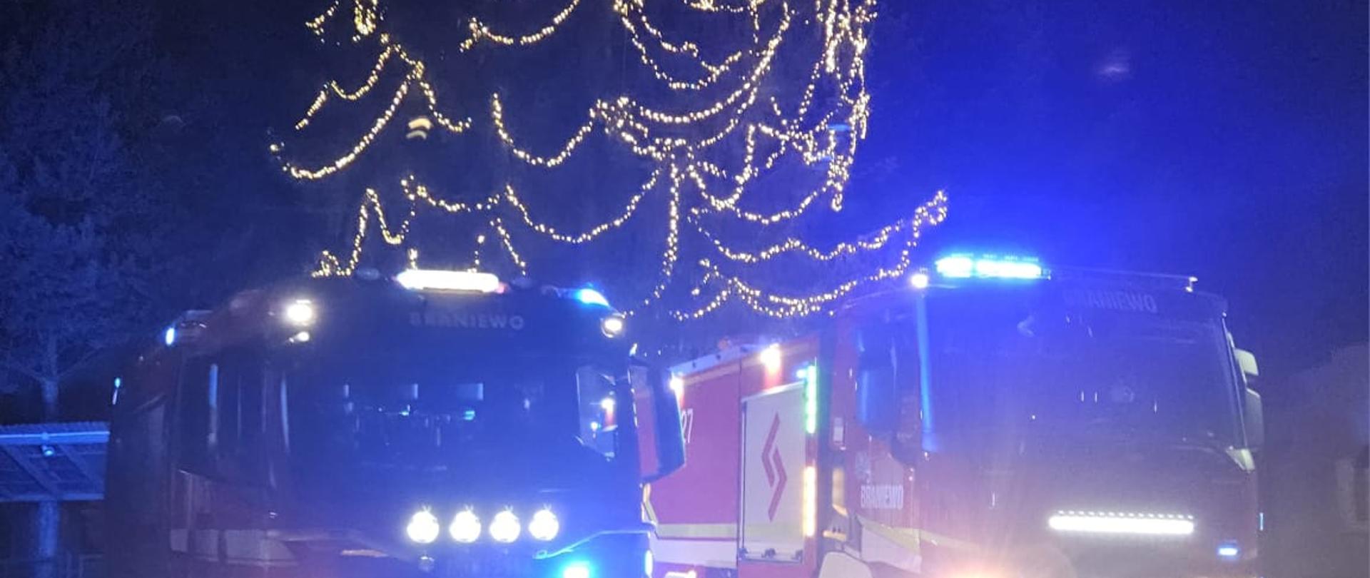 Zdjęcie zrobione nocą. Dwa duże samochody strażackie na światłach. W tle duża choinka udekorowana białymi lampkami.