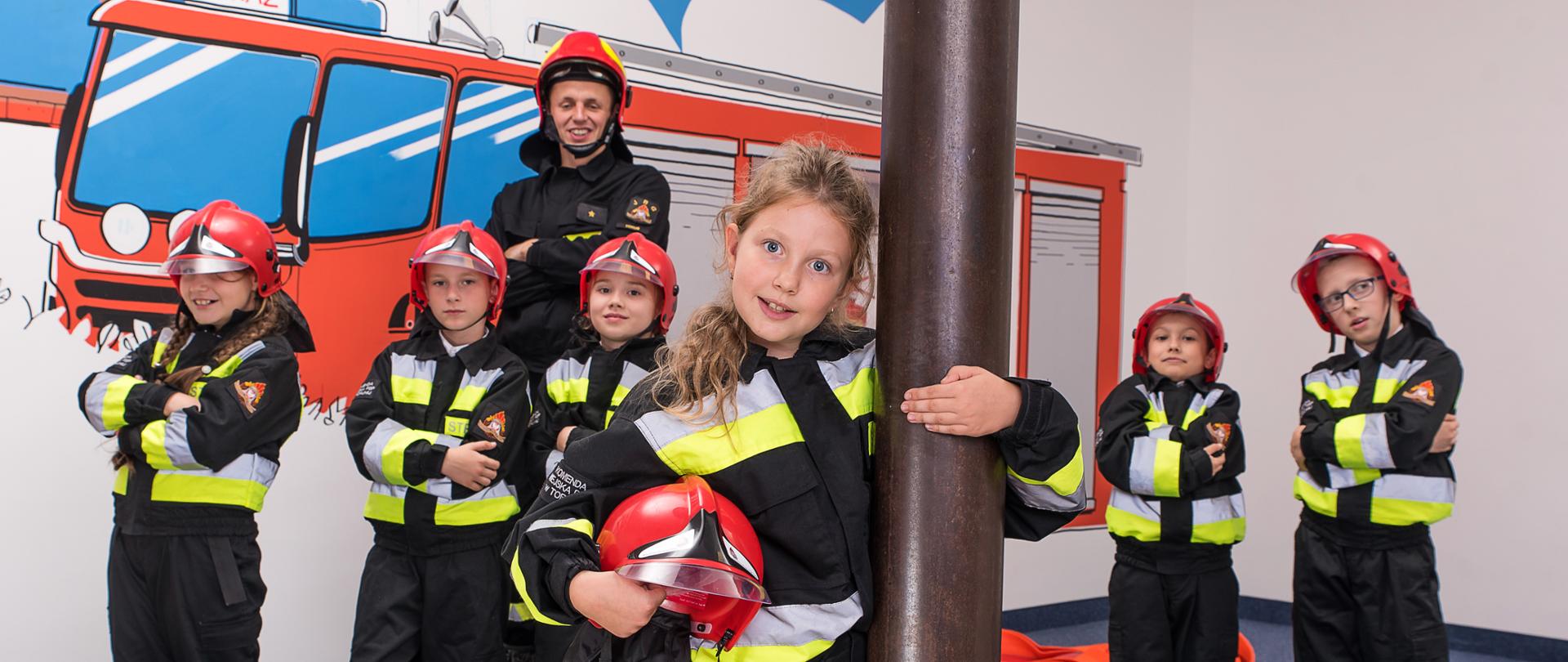 Na zdjęciu przy ześlizgu dziewczynka, za nią pięcioro dzieci i strażak. Wszyscy w mundurach hełmach. W tle na ścianie namalowany samochód strażacki.