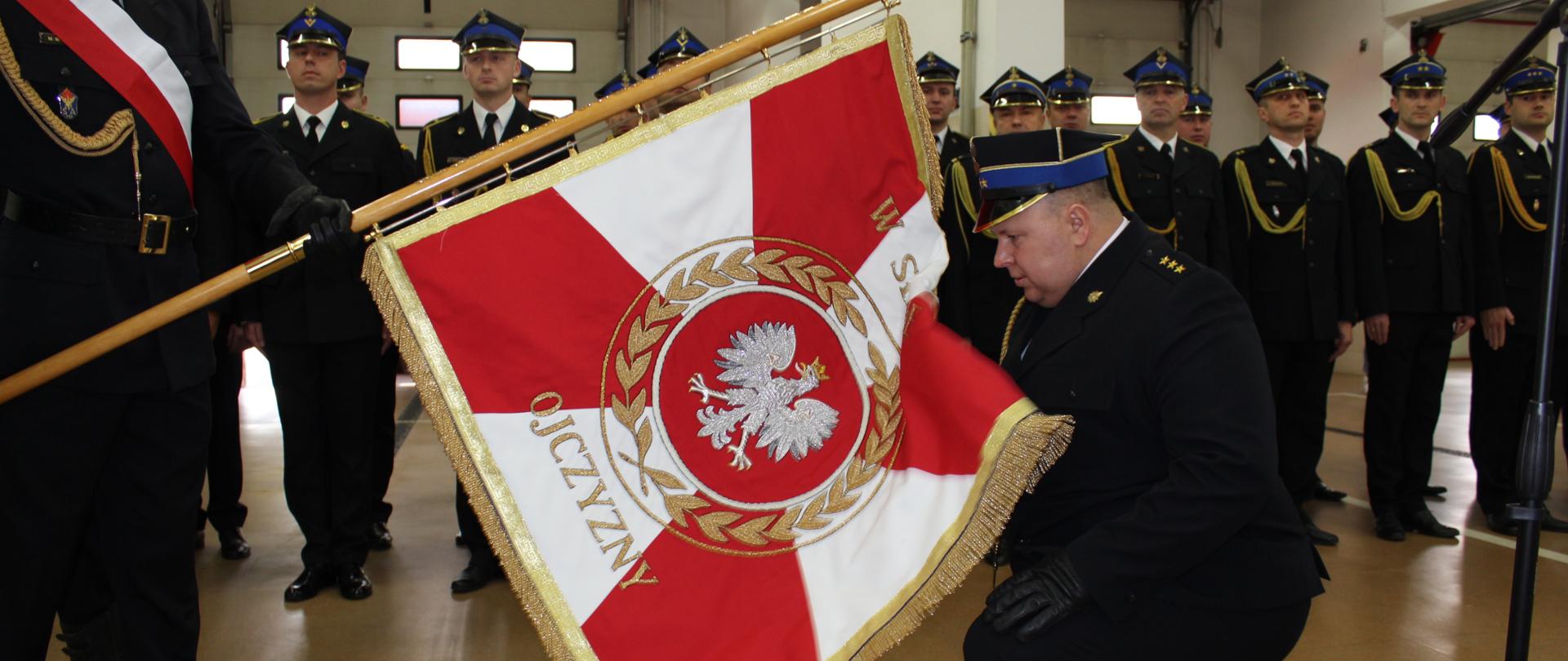 Komendant Miejski Państwowej Straży Pożarnej klęka na prawym kolanie przed pocztem sztandarowym, jednocześnie całując jego płat.