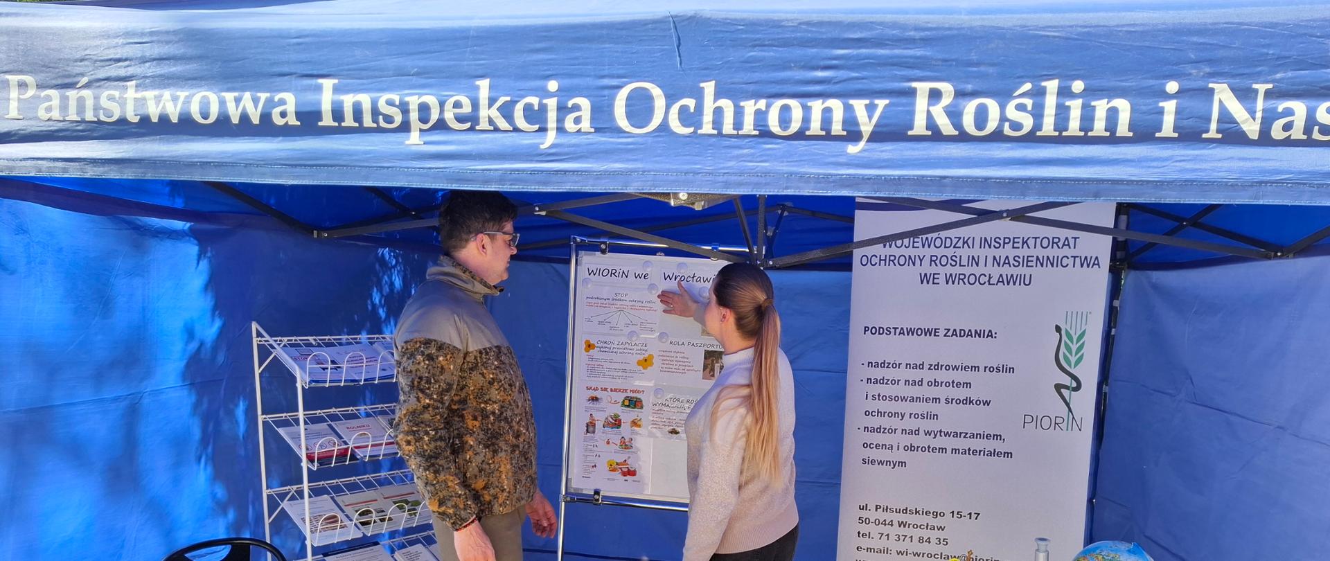 Mężczyzna stojący wewnątrz stoiska WIORiN w formie niebieskiego namiotu wystawowego. Obok kierownik Oddziału pokazująca na tablicy materiały informacyjne.