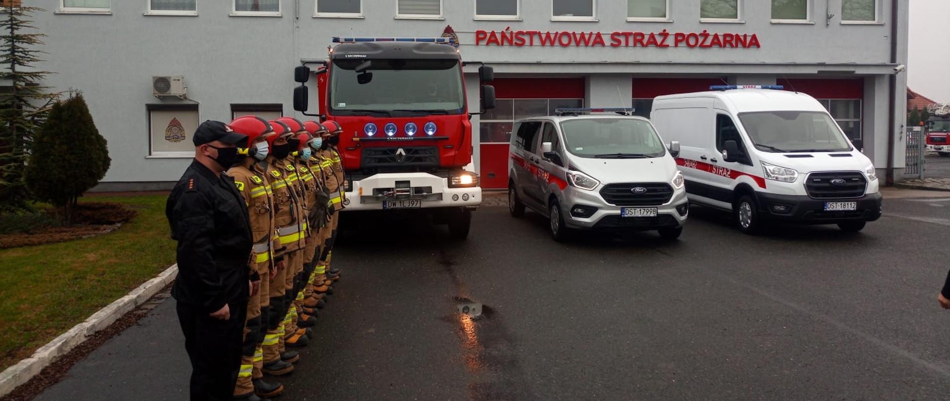 Zmiana służbowa stoi na baczność na tle trzech nowo otrzymanych pojazdów i budynku jednostki