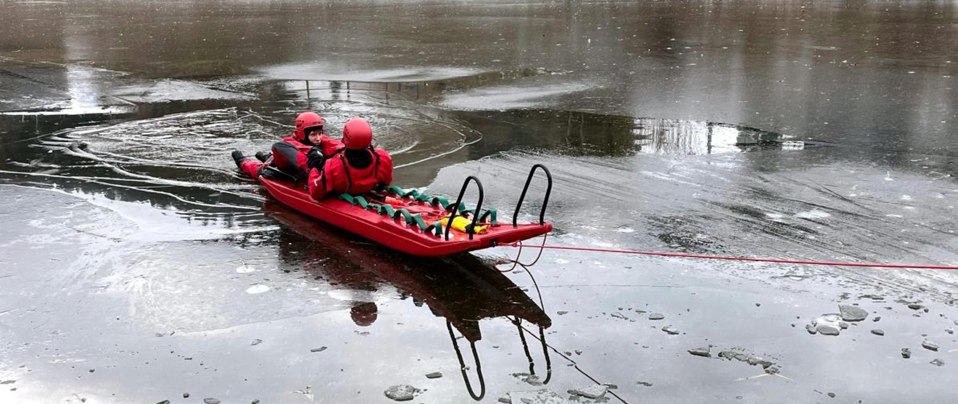 Na zdjęciu widać strażaków w skafandrze suchym na akwenie wodnym który pokryty jest lodem. Jeden ze strażaków znajduje się w wodzie a drugi podejmuje go z wody siedząc na saniach lodowych.