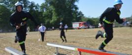 Zawody sportowo-pożarnicze w Jeziorach. Plac ćwiczeń. Trwa jedna z konkurencji zawodów - sztafeta pożarnicza 7x50 m z przeszkodami. Dwóch zawodników trzyających w ręce prądownice, biegnie po równoważniach, które mają kolor biało-czerwony. W tle drzewa, przeszkody i kibice.