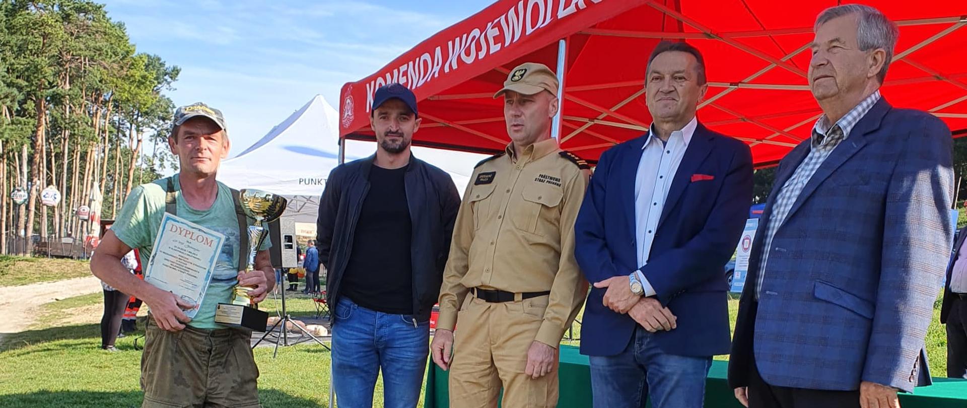 Zdjęcie obrazuje 4 osoby. W środku znajduje się zastępca świętokrzyskiego komenda wojewódzkiego PSP w Kielcach st. bryg. Grzegorz Rajca w umundurowaniu służbowym. Pierwszy mężczyzna od lewej trzyma w rękach puchar oraz pamiątkowy dyplom.