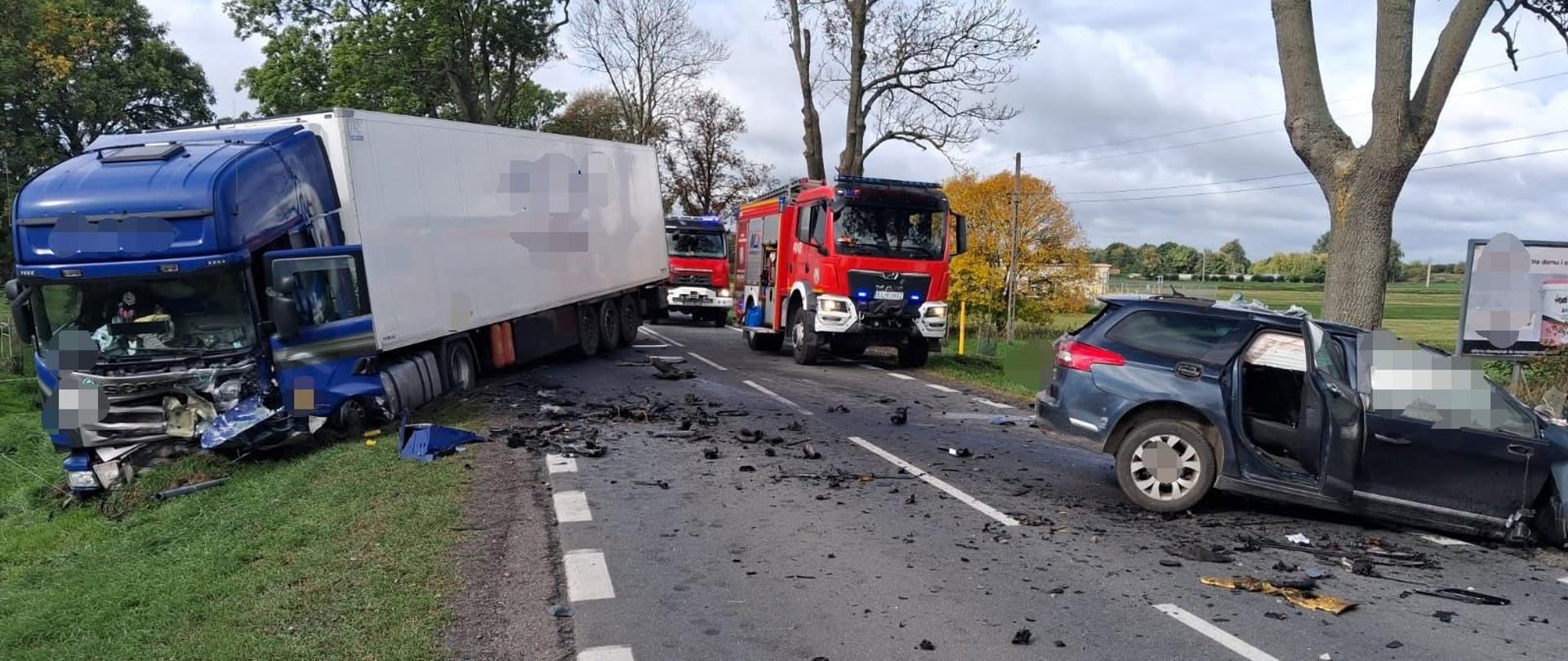 Na zdjęciu przedstawiono miejsce wypadku drogowego. Na drodze znajdują się dwa mocno uszkodzone pojazdy: po lewej stronie niebieska ciężarówka z naczepą, a po prawej ciemny samochód osobowy z otwartymi drzwiami. Na jezdni leżą liczne fragmenty pojazdów i inne części, które zostały rozrzucone w wyniku zderzenia. W tle widoczne są wozy straży pożarnej oraz drzewa i pola. 