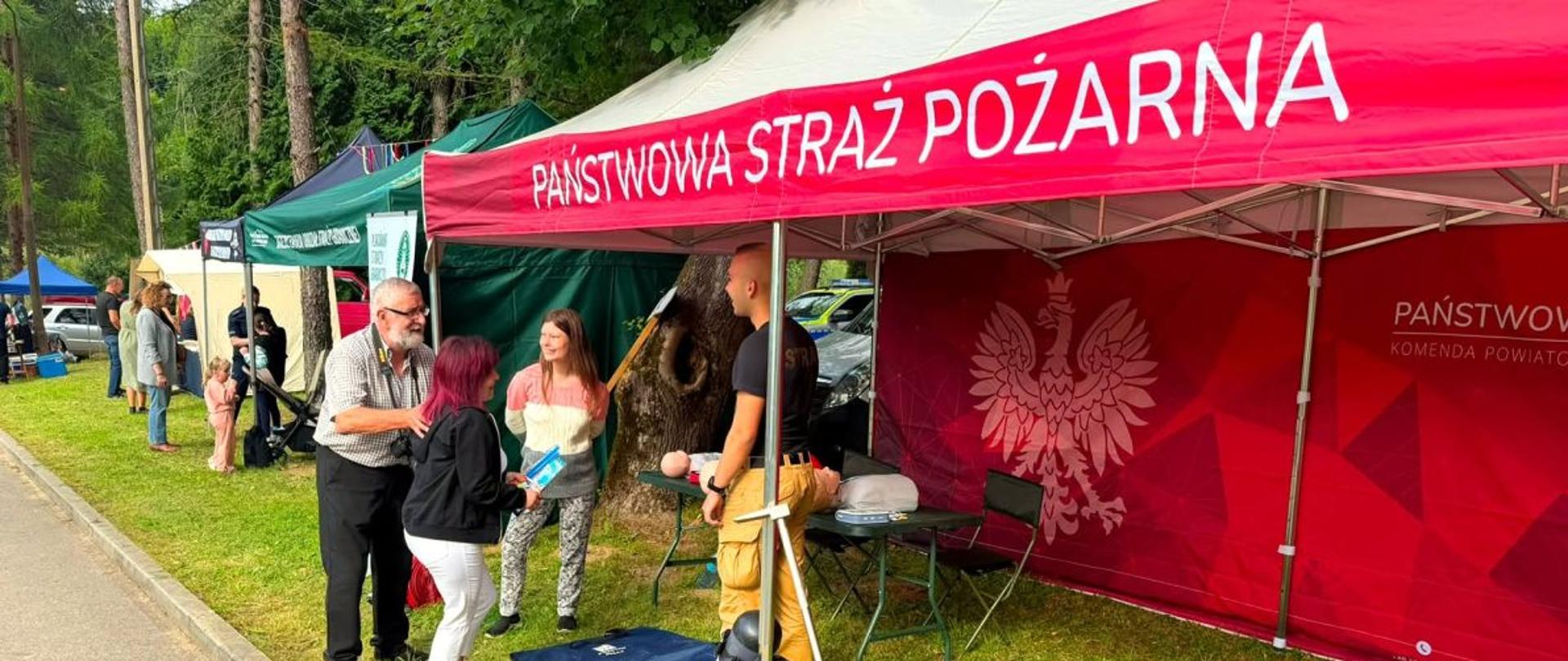 Zdjęcie przedstawia stoisko PSP na dożynkach. Przy stoisku stoi strażak oraz zainteresowane osoby. 